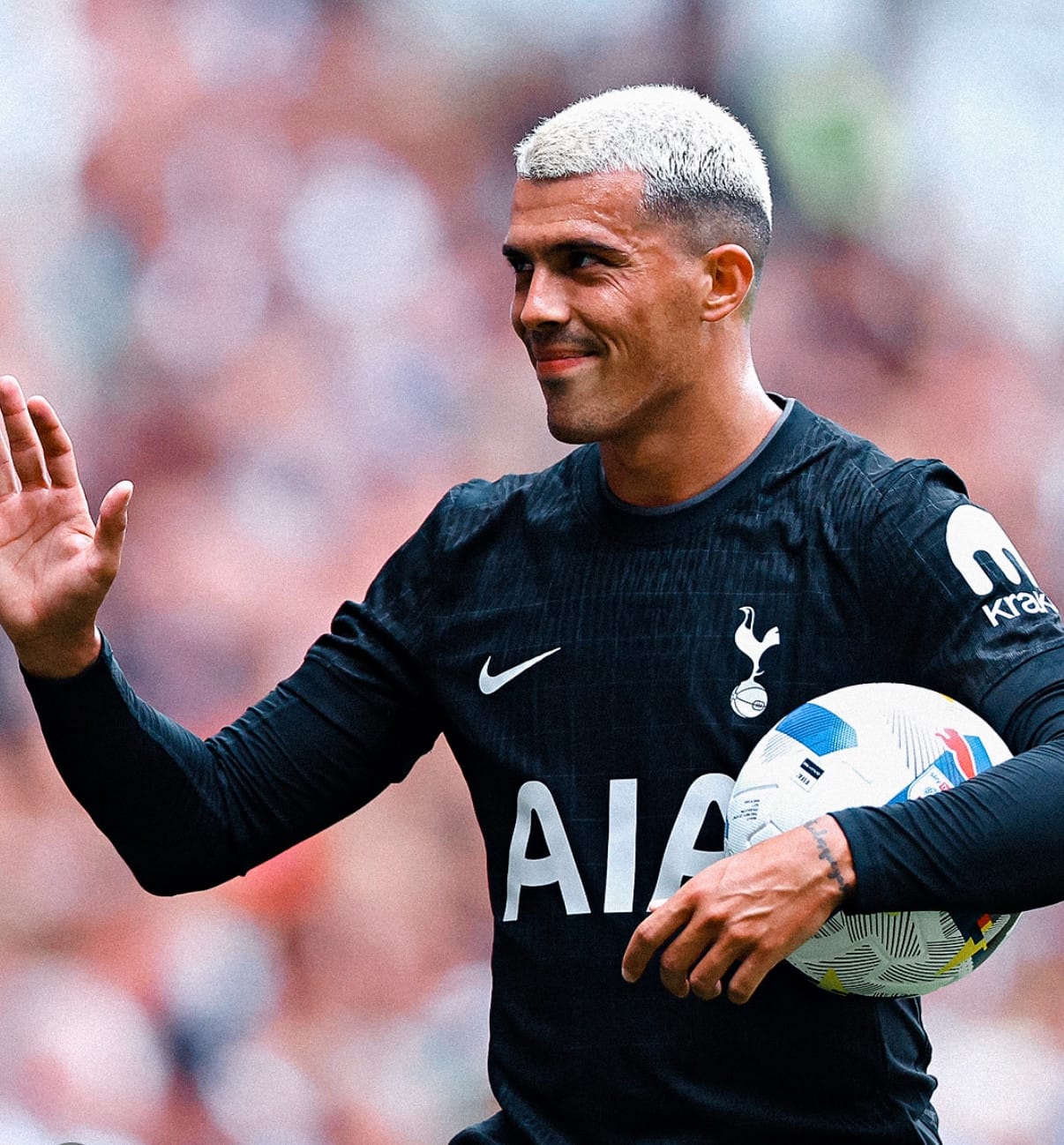 A smiling Pedro Porro, with blonde hair and wearing the black away kit, holds the ball in one hand and waves with the other.