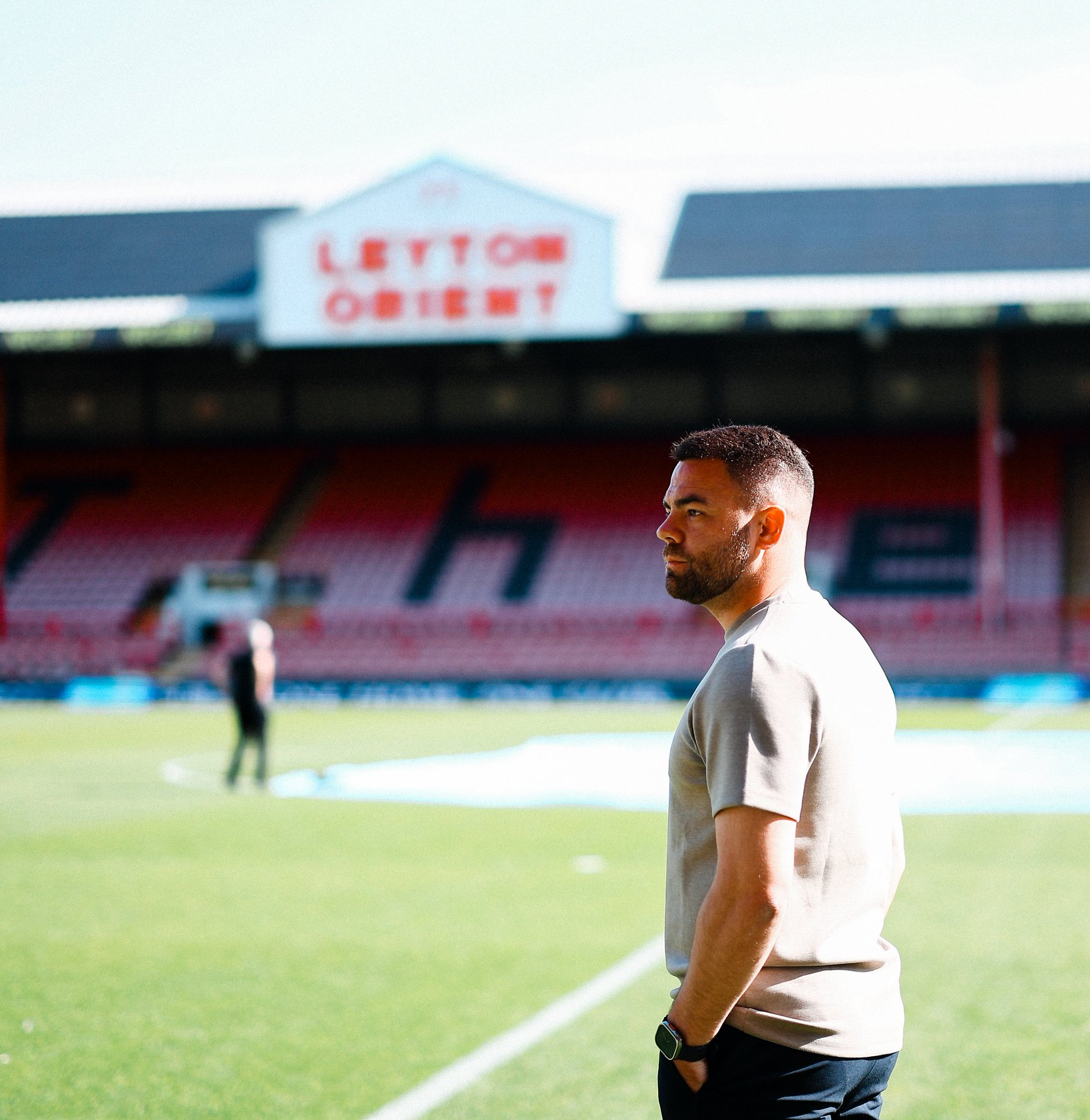 Martin Ho, hands in pockets, takes in the environment at Brisbane Road, with the Leyton Orient sign visible but blurred in the background.