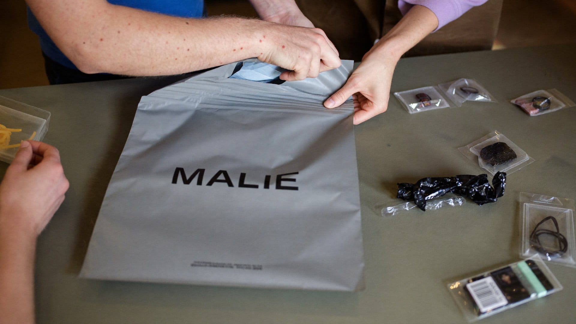 Hands opening a grey poly mailer with inexpensive goods on a table