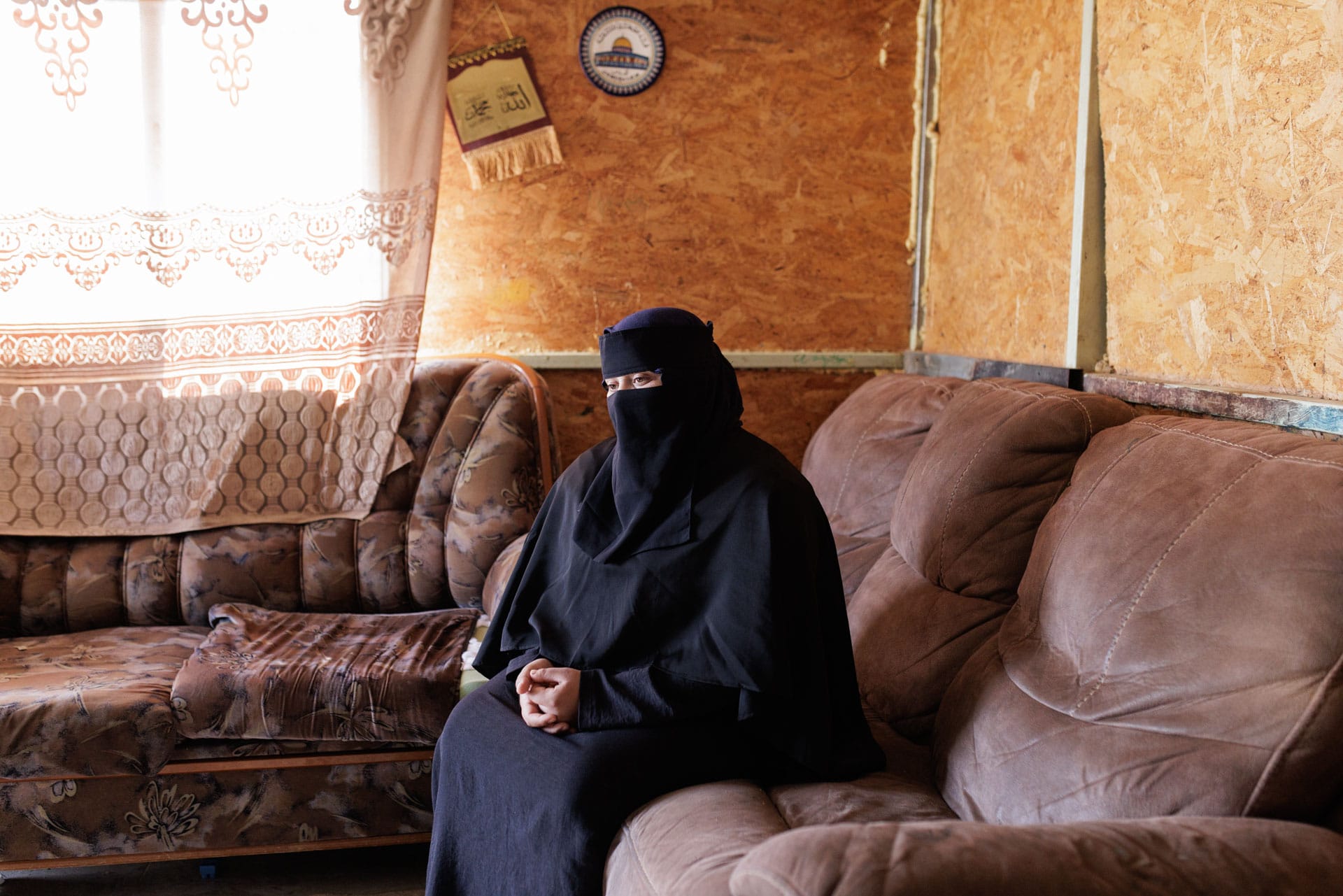 A woman wearing a niqab sits on a sofa in a sparsely furnished room, with light coming through a window to her left.