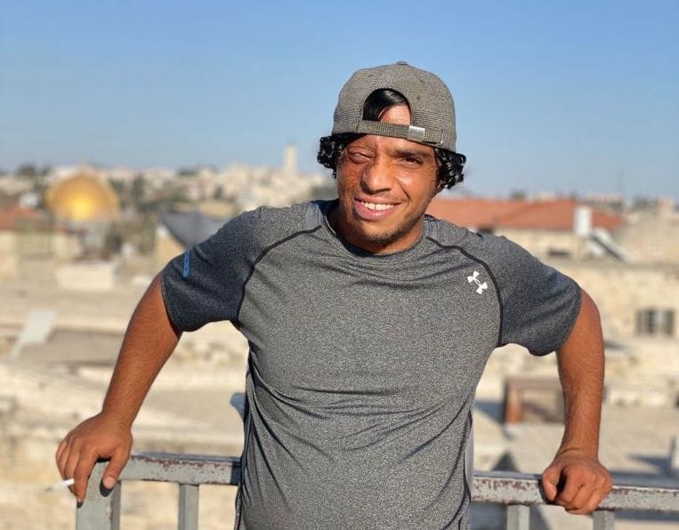 A man wearing a black cap and dark T-shirt stands at a viewpoint overlooking a tree-lined road and city buildings under a clear blue sky.