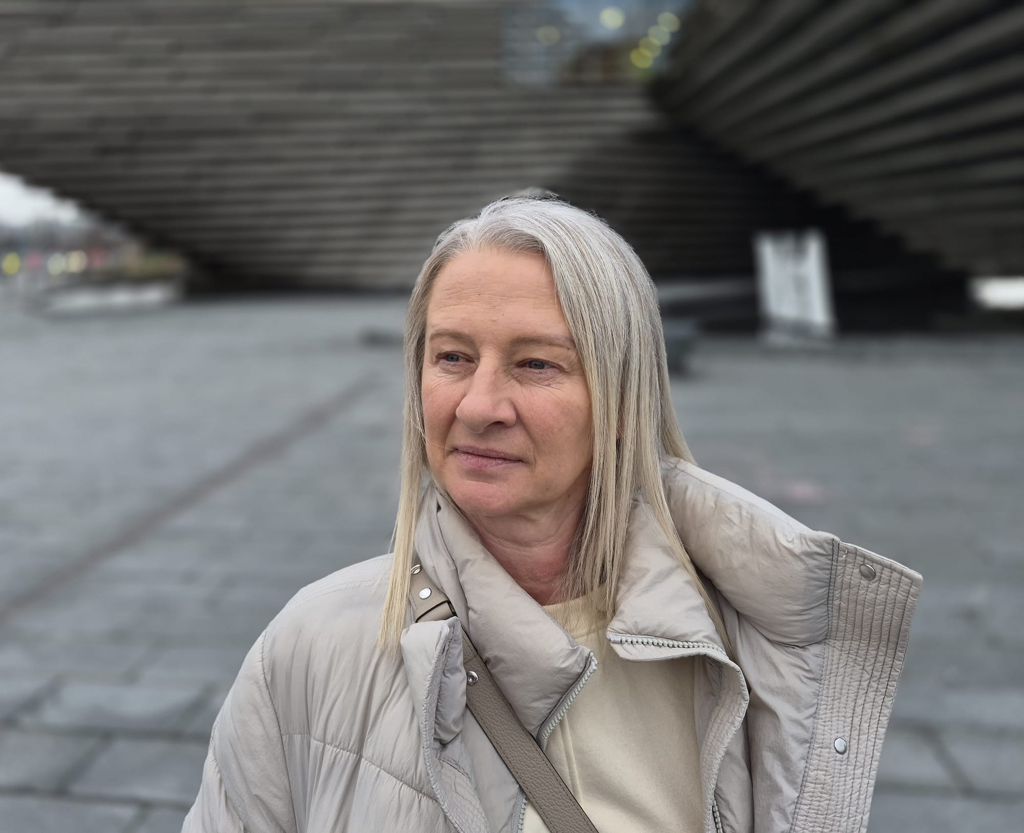 Middle-aged woman with long grey hair standing outdoors in an urban square, with modern buildings blurred in the background.