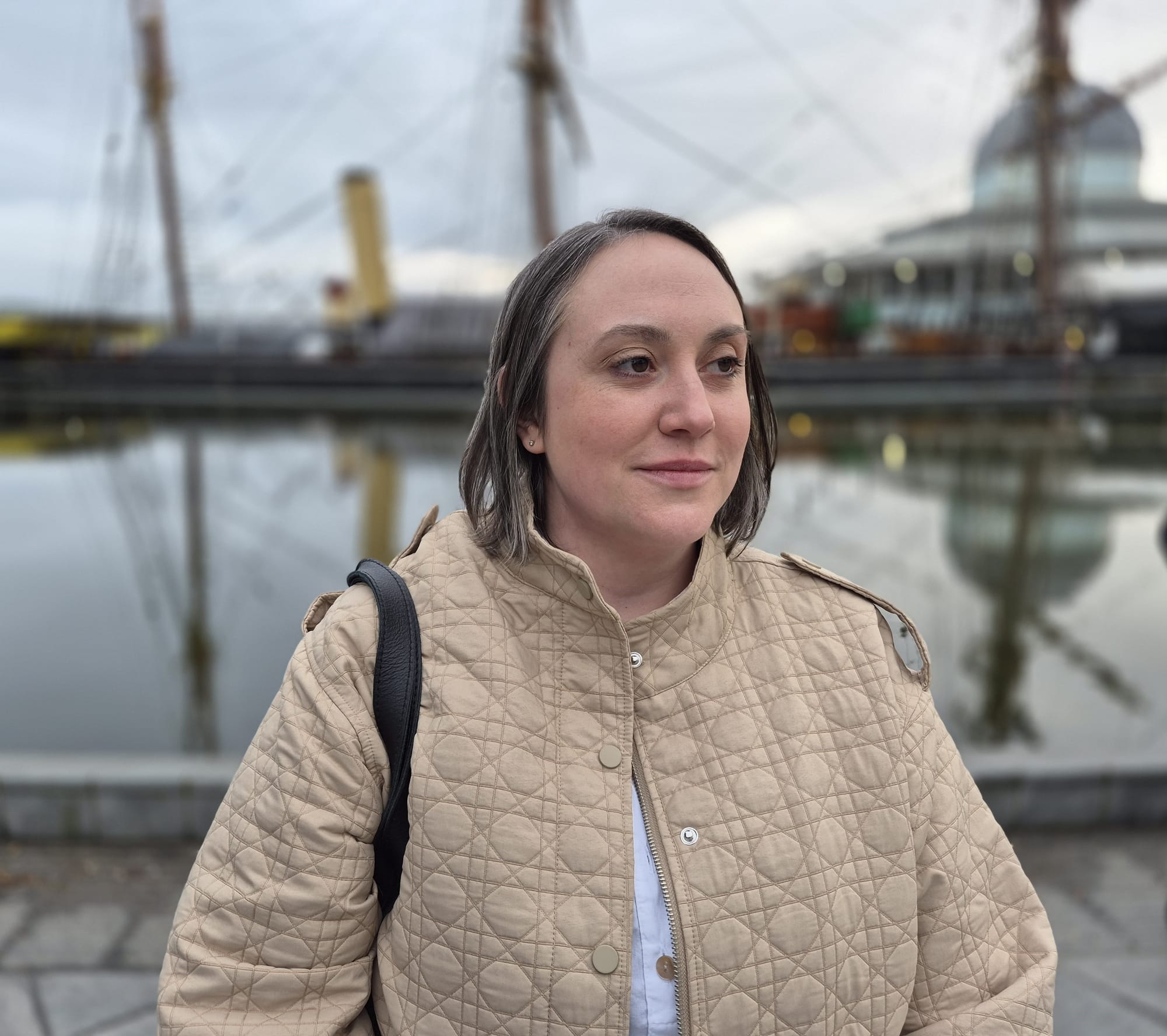 Woman in a beige jacket standing by a waterfront, with boats and buildings reflected in calm water behind her.