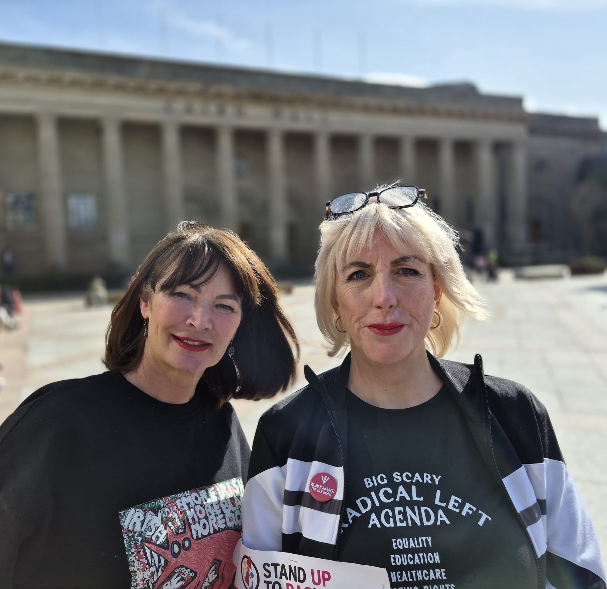 Two women standing outdoors in front of a large building, wearing T-shirts with political slogans.