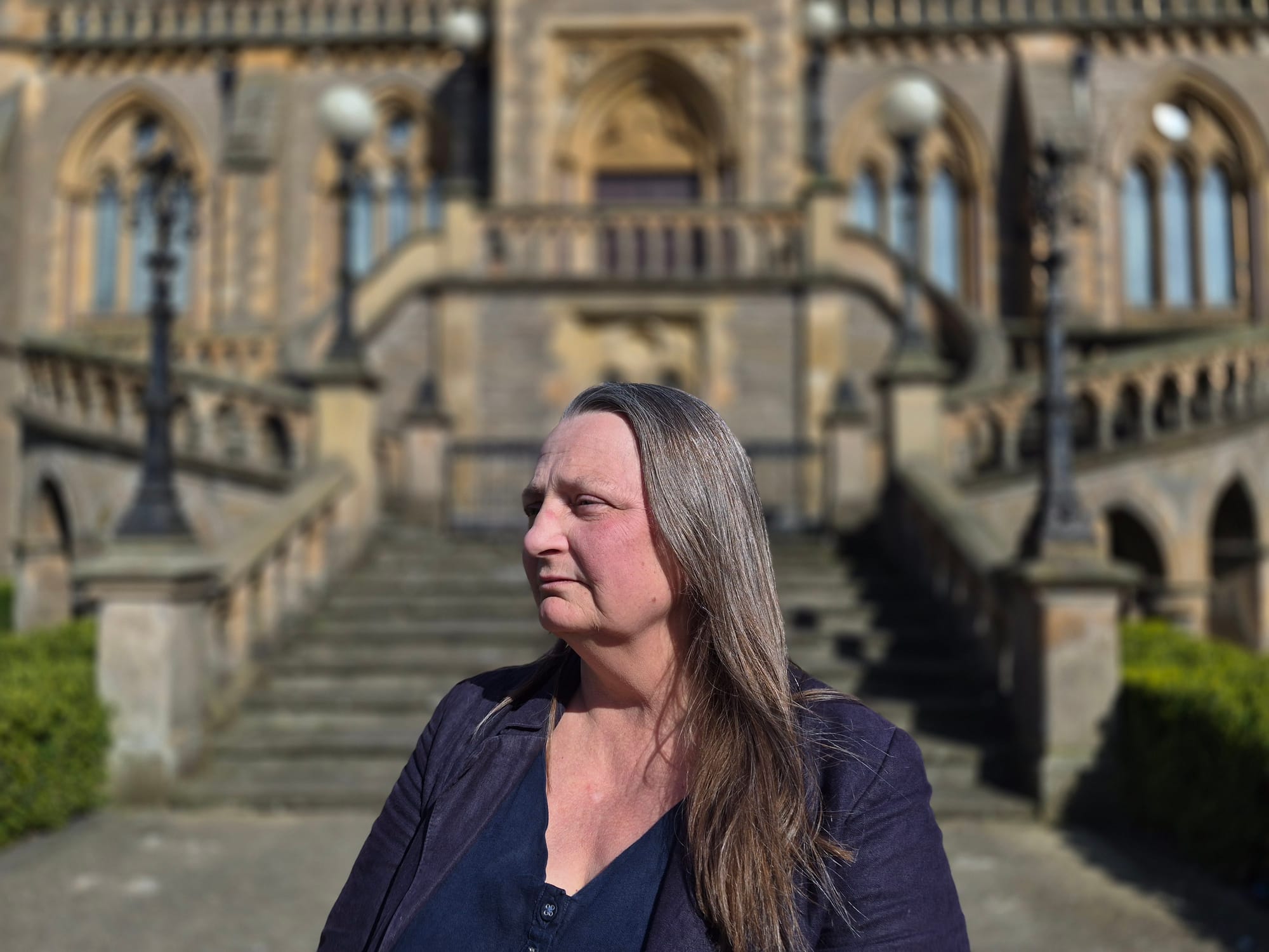 Woman standing in front of a large historic building with stone steps and arches, looking to the side.