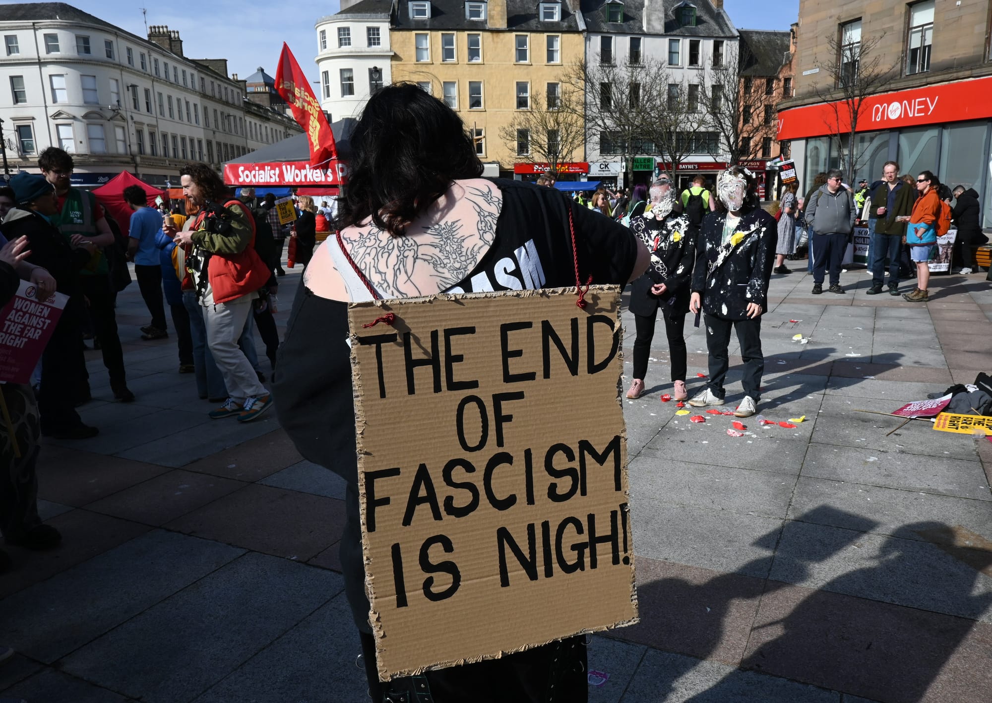 Person wearing a cardboard sign reading “The end of fascism is nigh!” at a busy outdoor protest.