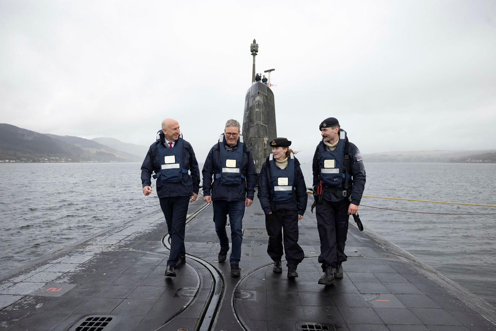 Four people, including a uniformed naval officer, walking along the deck of a surfaced submarine in a Scottish loch, wearing life jackets with hills and water in the background.