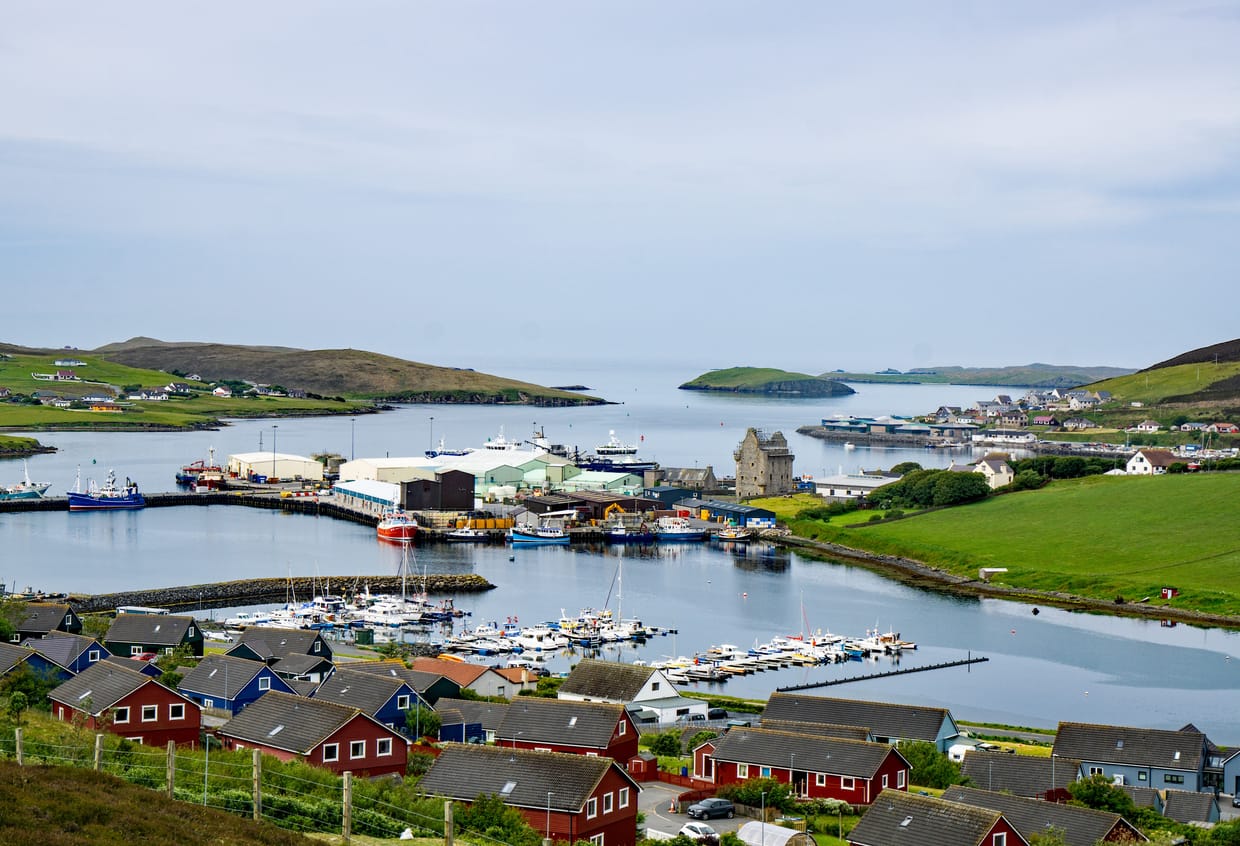 A coastal town and harbour in Shetland, with boats moored in calm water, red-roofed houses in the foreground, and green hills and islands in the distance.