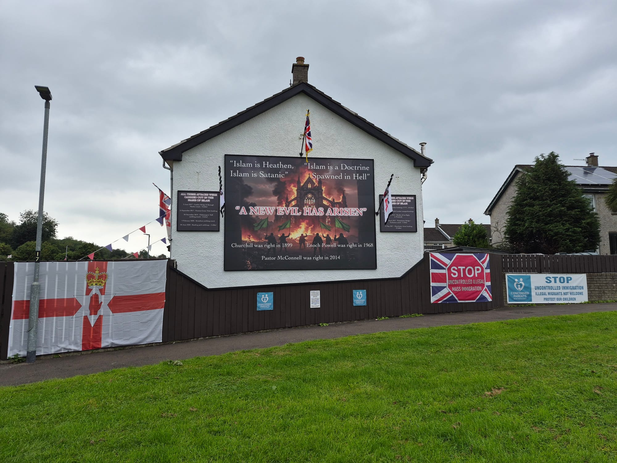 Exterior of a building decorated with Union Jack and Northern Ireland flags, anti-immigration banners and a large anti-Islam sign reading “A new evil has arisen.”