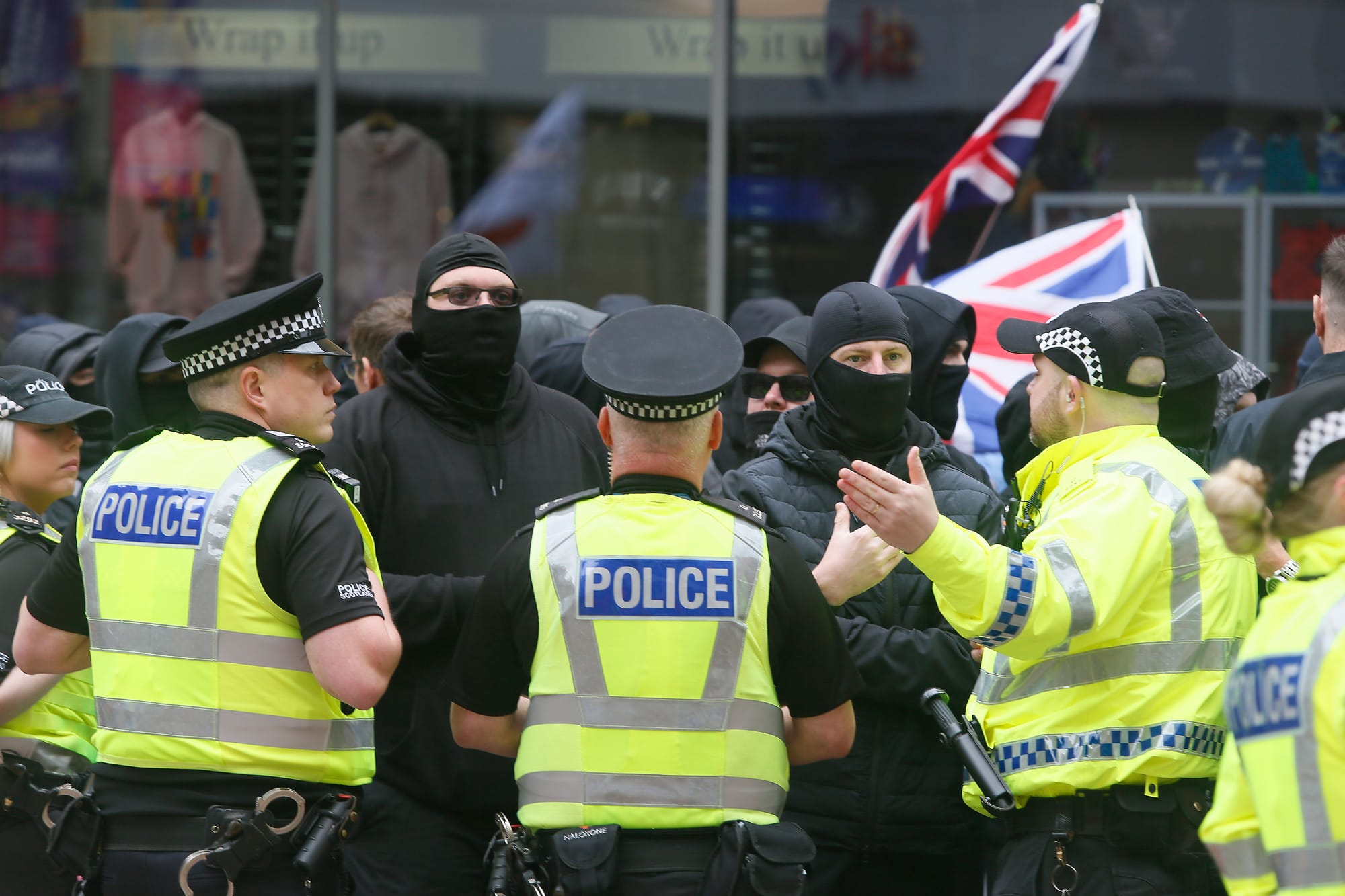 Police officers stand face-to-face with a group of masked protesters dressed in black, with Union Jack flags visible in the background.