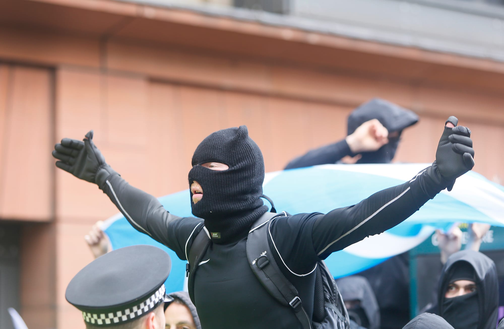 A masked protester dressed in black stands with both arms outstretched in front of a Saltire, with police and other masked demonstrators around him.