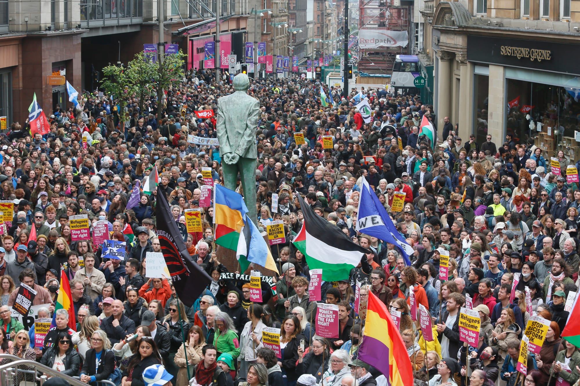A large counter-protest fills a Glasgow city street, with crowds carrying placards and Palestinian, Ukrainian, Pride and other flags around a central statue.