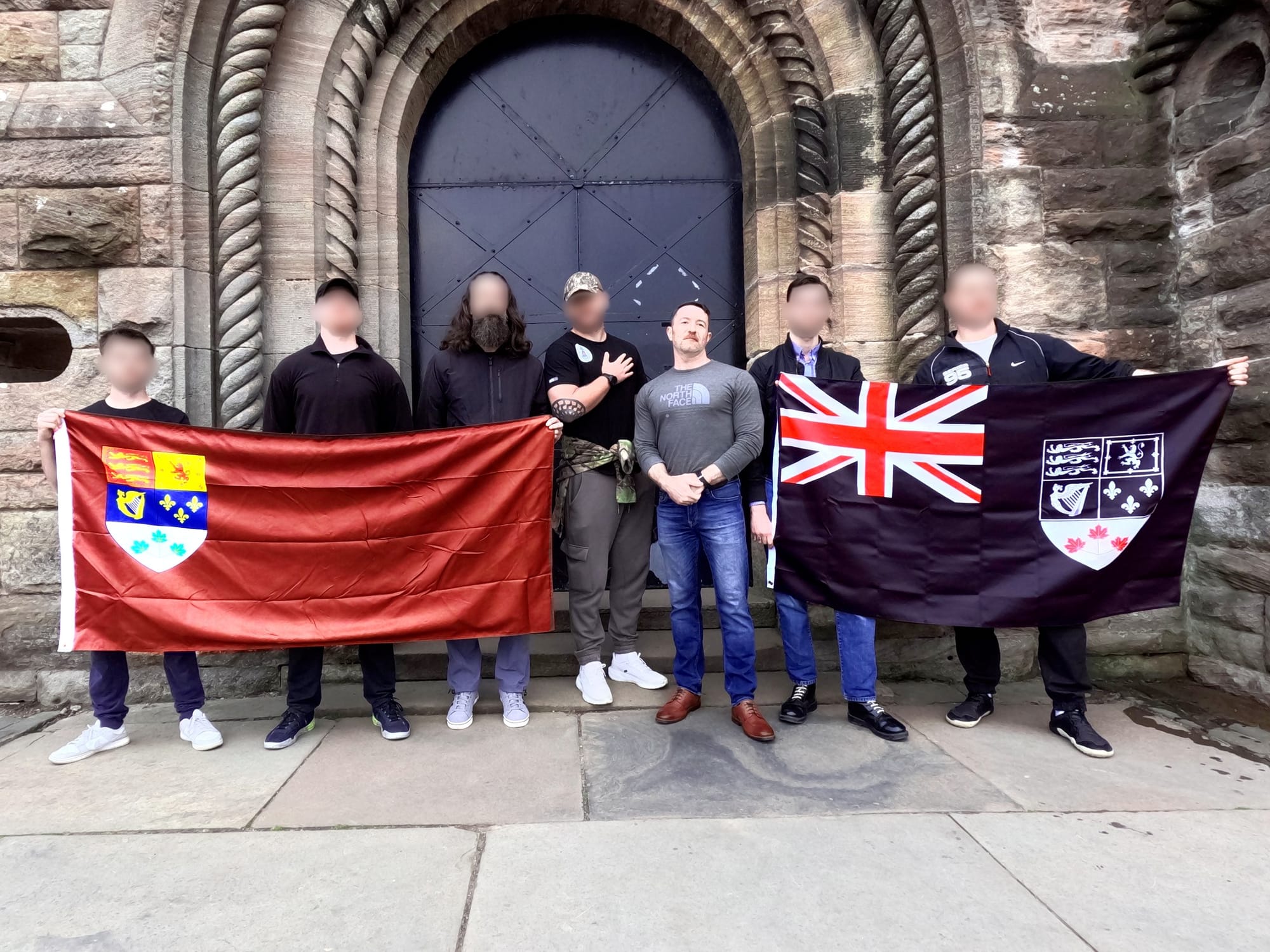 A group of people, most with faces blurred, stand outside a stone building holding two large flags featuring heraldic crests. A man with an unblurred face stands in the centre.
