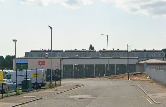 Industrial building at the end of a quiet access road, with fencing, streetlights and several vehicles parked outside.