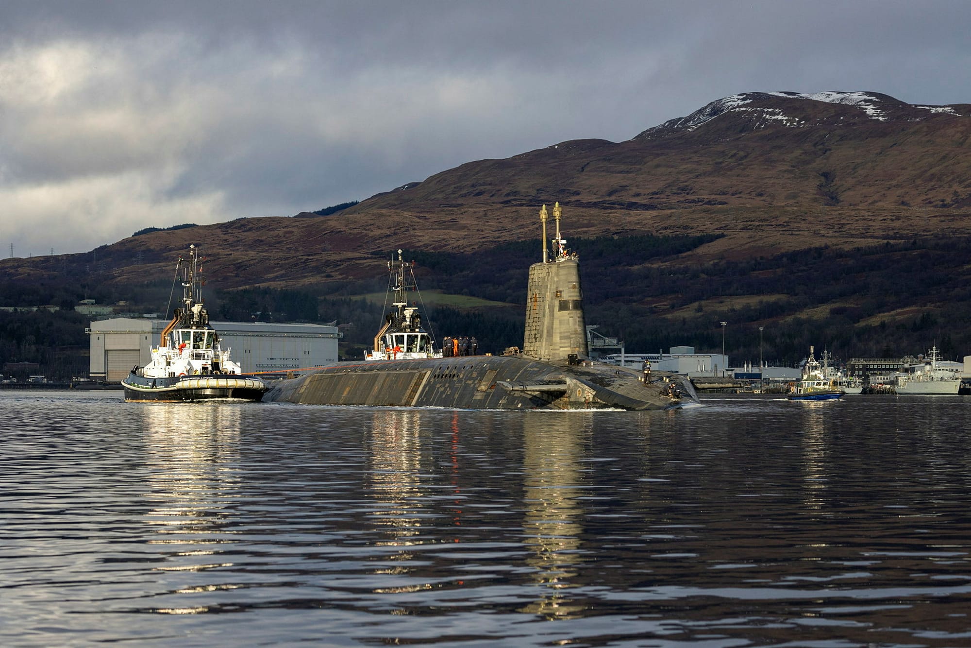 Royal Navy nuclear submarine HMS Vengeance on the Gareloch near Faslane, accompanied by tugboats, with hills rising behind the naval base.