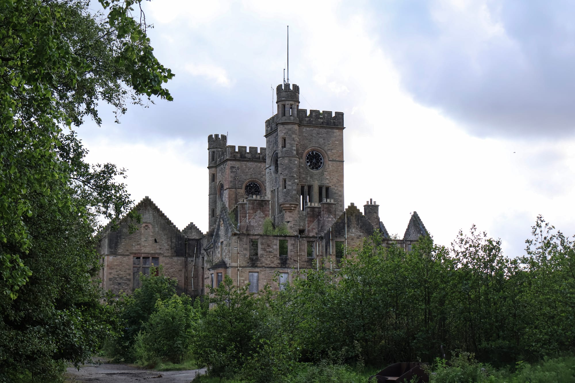 The abandoned nurses’ home at Hartwood Hospital, seen through trees and overgrown grounds, with many windows broken or boarded and a security fence across the front.
