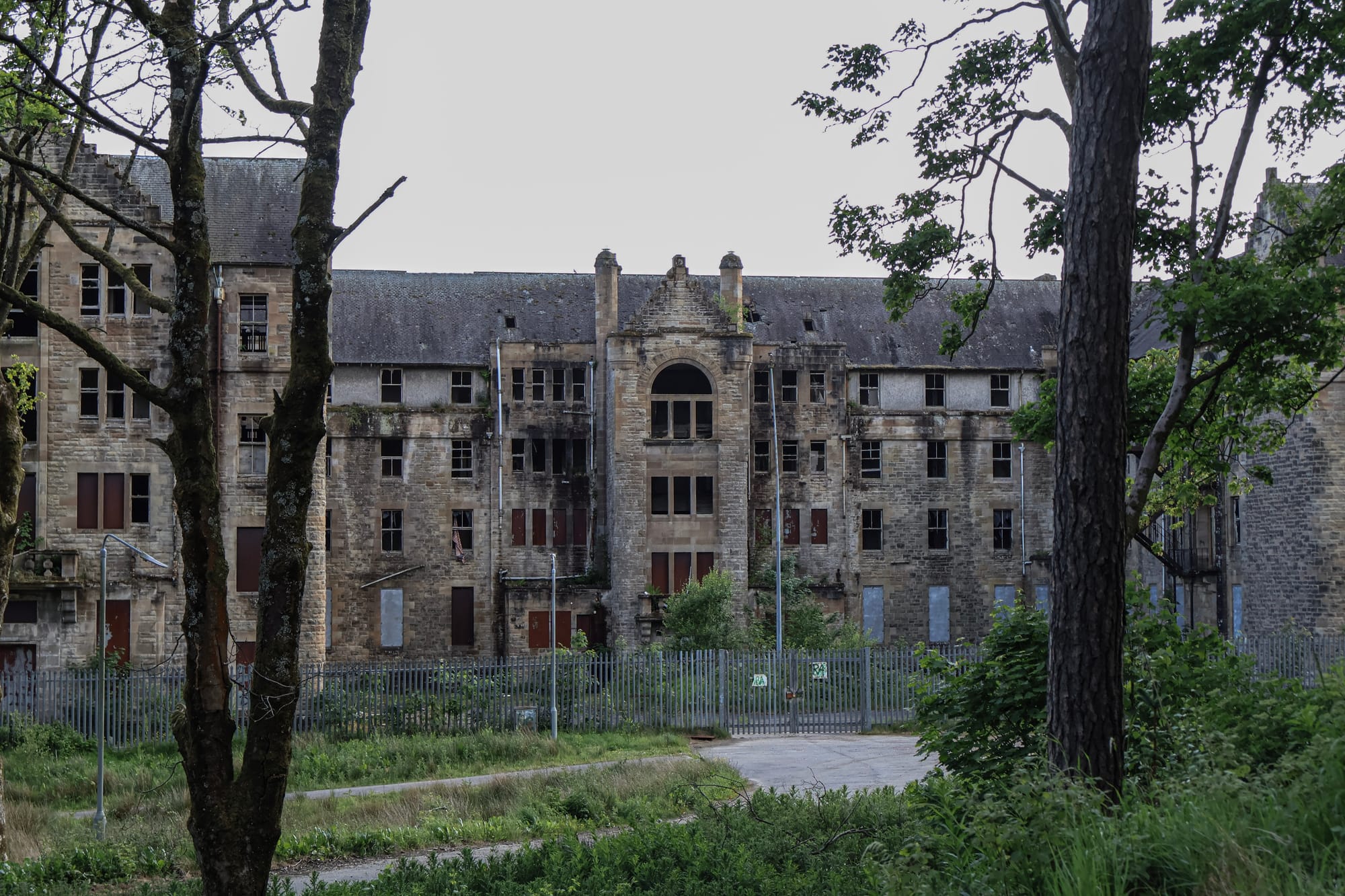 Hartwood Hospital viewed through overgrown trees, showing its ruined stone buildings and two castellated clock towers beneath a cloudy sky.