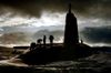 Silhouetted crew members stand and move across the hull of a surfaced military submarine at sea, with clouds overhead and waves breaking around the vessel as light breaks through the sky.