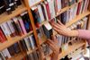 A close-up of a person’s hands removing a book from a wooden bookshelf filled with tightly packed books in a library.