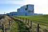 Torness nuclear power station, a large white industrial complex, stands beside green farmland and a fenced path near the Scottish coastline under a blue sky.