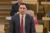 Anas Sarwar, leader of Scottish Labour, stands speaking in the Scottish Parliament chamber, wearing a dark suit and red tie. Other MSPs sit behind him at desks, listening.