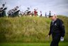 A man in golf attire walks across a grassy course while a group of bagpipers play in the background.