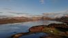 Aerial view of a coastal Scottish loch with calm water, fish farm cages offshore, and hills and mountains in the background under a clear sky.