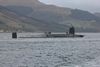 Royal Navy submarine on the surface in a Scottish loch, with crew members standing on deck and hills and shoreline in the background.