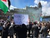 A large crowd gathers in Glasgow during a pro-Palestine protest, with people holding flags and placards. In the foreground, a person holds a handwritten sign reading “HANDS OFF PALESTINE”.