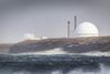 Coastal industrial site with a large white domed structure and chimneys on a rocky shoreline, waves breaking in the foreground under a grey sky.