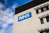 Exterior of an NHS building with a large blue and white NHS logo mounted on the cream-coloured wall beneath a blue sky with scattered clouds.