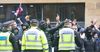 Police officers in high-visibility vests face a crowd of mostly masked protesters, some raising their arms, with a Union Jack flying behind them.
