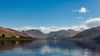 A calm loch surrounded by rugged mountains under a bright blue sky with scattered clouds, with a few houses visible along the shoreline.