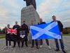 1. A group of people, most with faces blurred, pose beside the Robert the Bruce monument. They hold a Scottish Saltire and a black flag featuring the Union Jack and a heraldic crest.