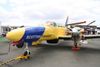 A yellow and blue Scottish Fisheries twin-engine aircraft is parked on display at an air show, with its propellers visible and people and other aircraft in the background.