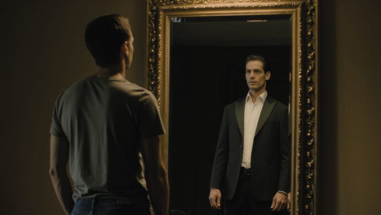 A man in a t-shirt faces a mirror, his reflection showing a formally dressed, confident figure — symbolising the Ego as a constructed self-image.