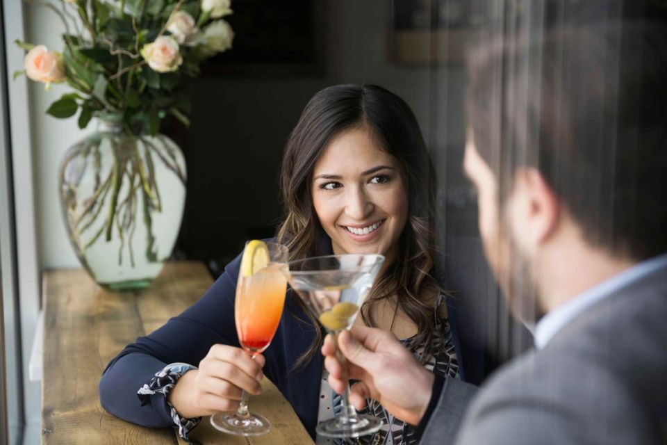 A woman in professional attire smiling warmly while clinking cocktail glasses with a man whose back is to the camera. An intimate moment of genuine connection.