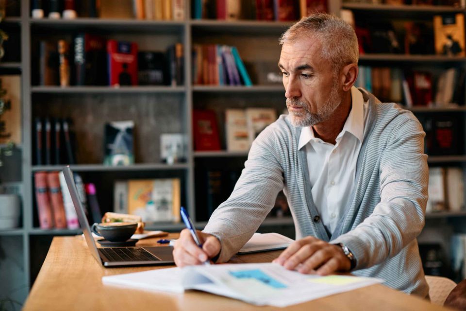 A mature silver-haired man in his fifties sitting at a wooden desk covered in books and notes, writing with focused concentration. 
