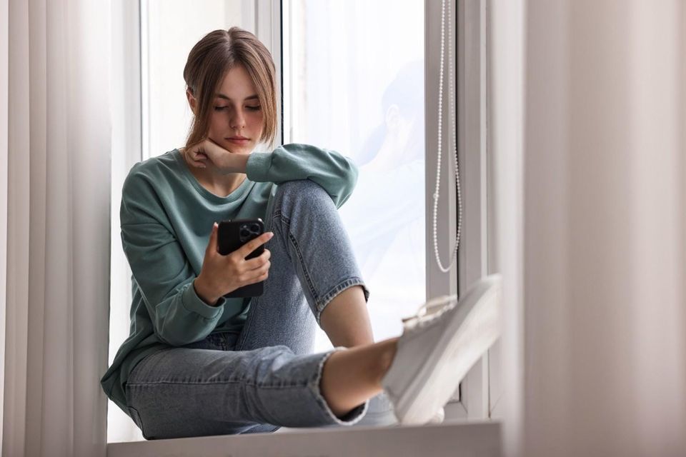 A young woman sits curled up on a window sill, looking down at her phone with a pensive, melancholy expression, her chin resting on her knee.