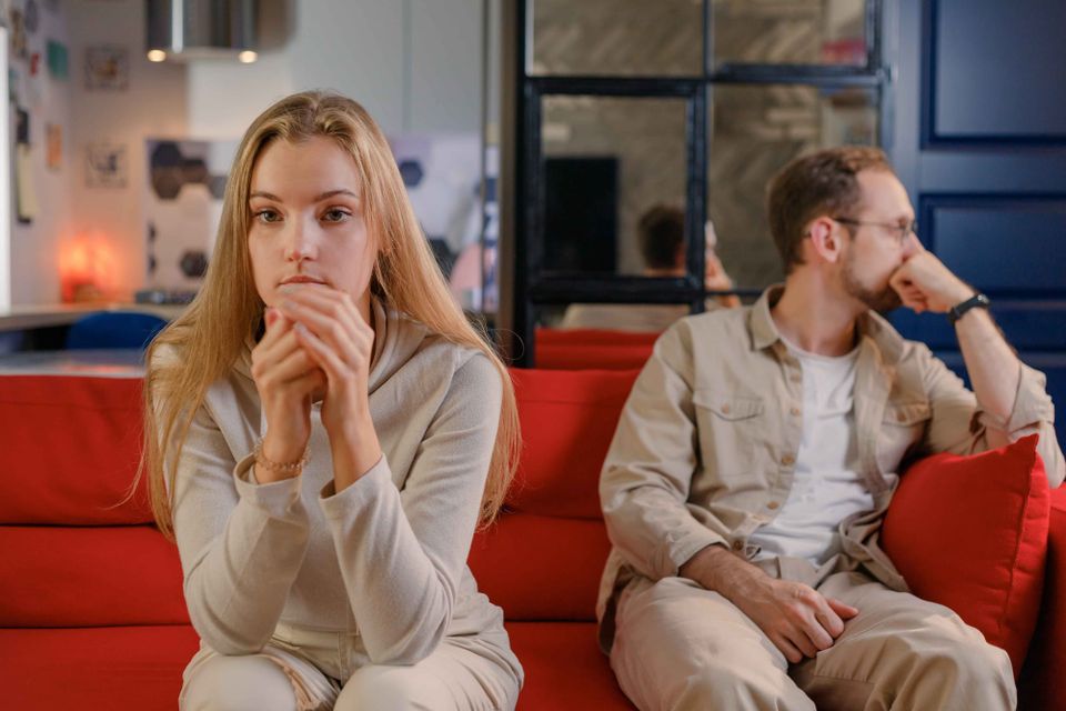 A young couple sitting apart on a red couch, both lost in silence. She looks forward with worry in her eyes, hands clasped. He sits behind her, head resting on his hand, looking away. 
