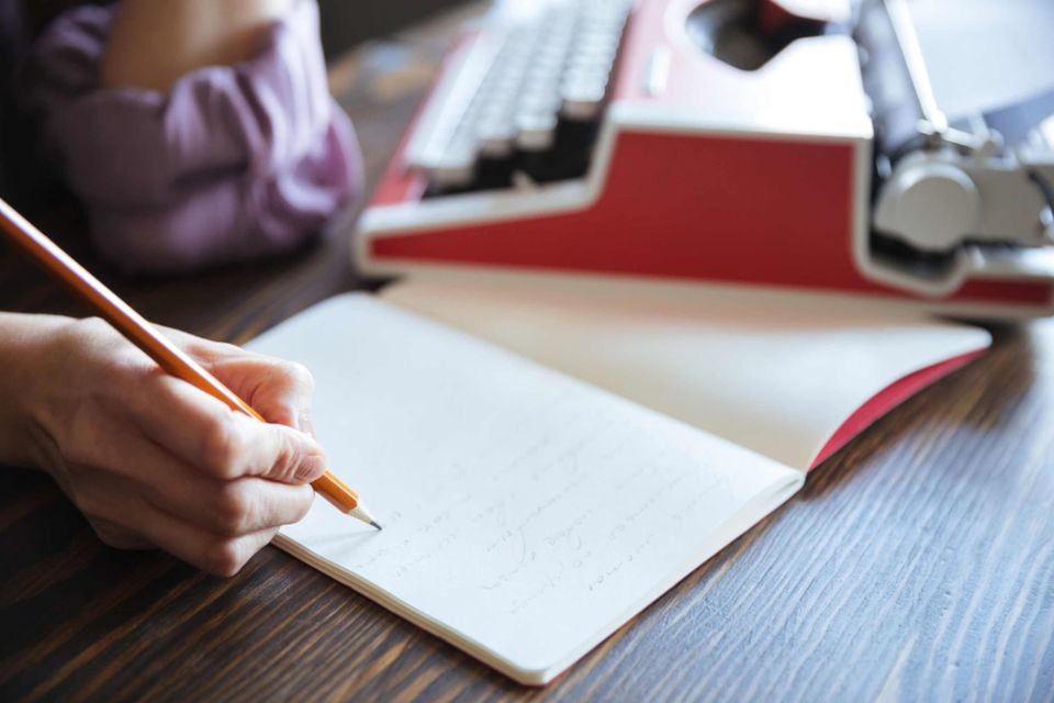 A hand holding a pencil over an open notebook, with a vintage red typewriter in the background on a wooden desk.