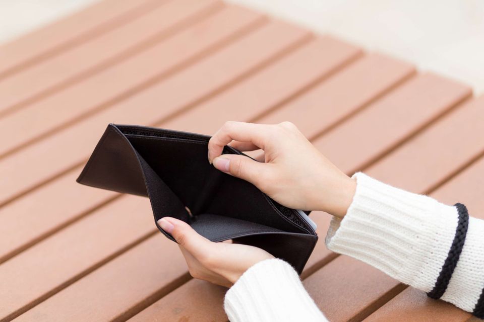  woman in a white knit sweater holds an open empty wallet over a wooden surface. The wallet is completely empty. 