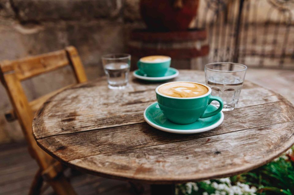 Two teal cappuccino cups and water glasses on a rustic wooden table. An empty chair sits in the background.