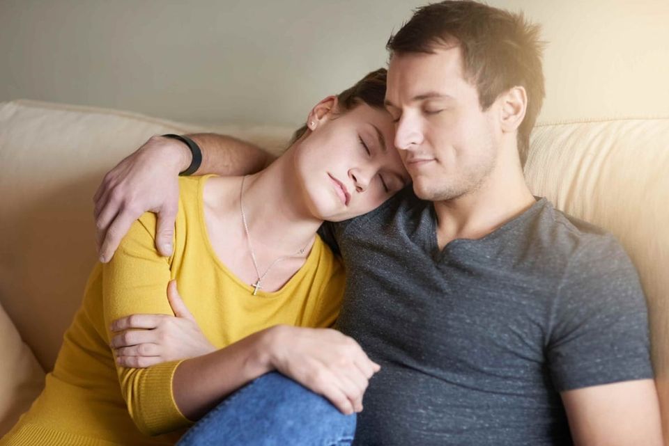 A couple rest together on a sofa, she leaning against him with eyes closed, his arm around her. The atmosphere is warm and tender — two people who have found their way back to each other.