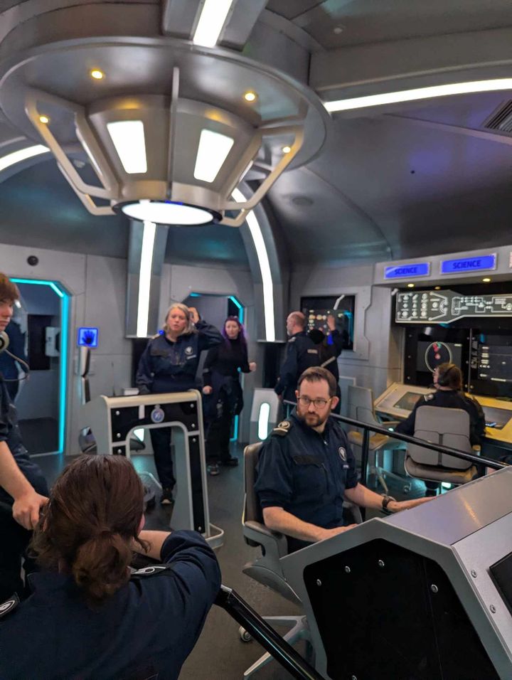 A group of people in blue jumpsuits on the set of a space ship, playing Bridge Command