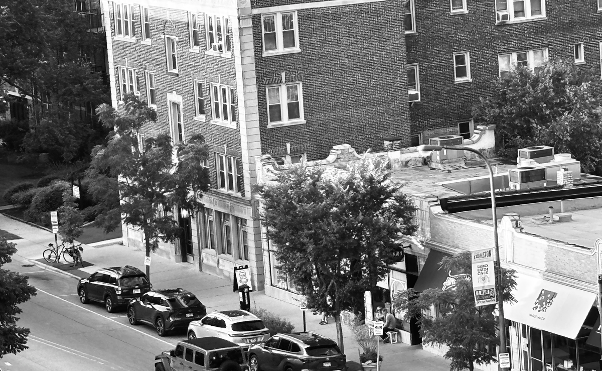 Apartment building, retail shops and cars parked along the street. Black and white.