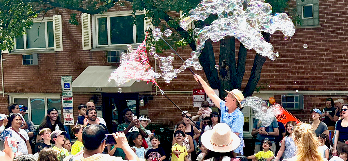 Man in a hat making big bubbles at a street fair.