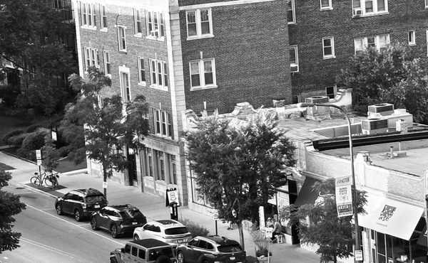 Apartment building, retail shops and cars parked along the street. Black and white.