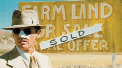 Un homme au nez bandé devant un panneau Farm land for sale barré du mot Sold