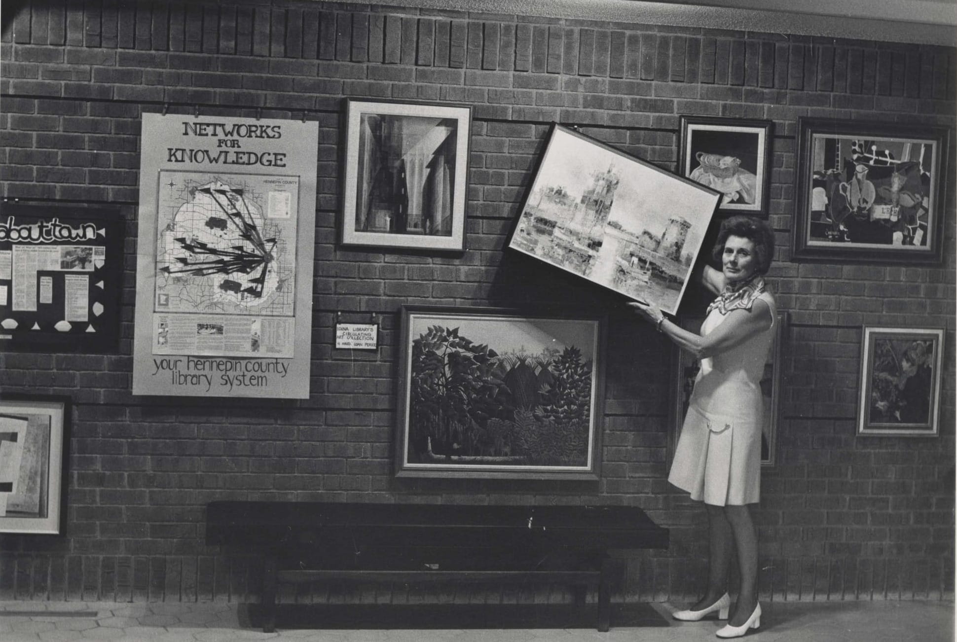 A woman holds a framed picture next to a wall of framed picture that are loaned to library patrons